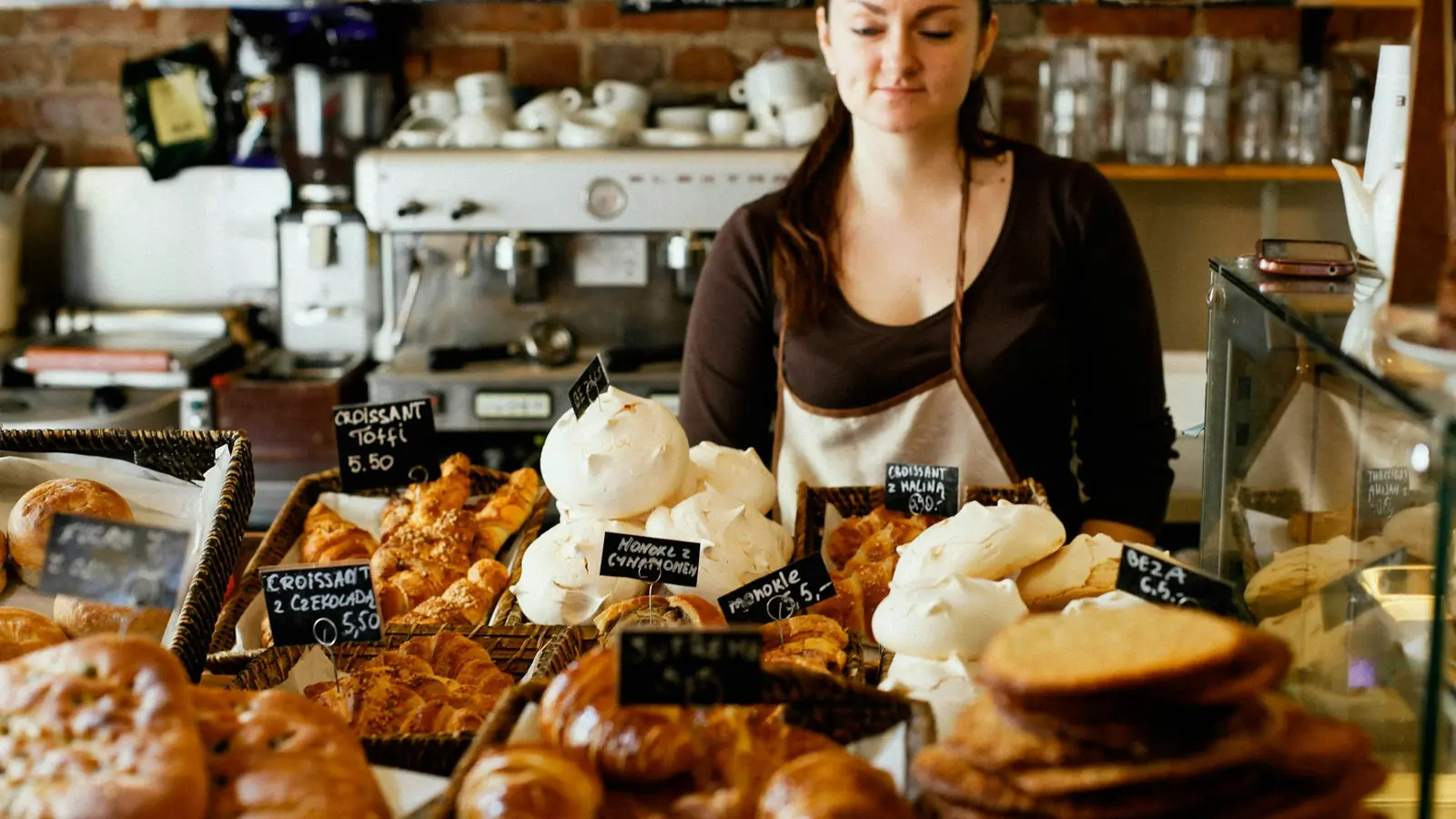Eine Frau mit Schürze steht hinter der Theke einer Bäckerei, vor ihr Croissants und Gebäck