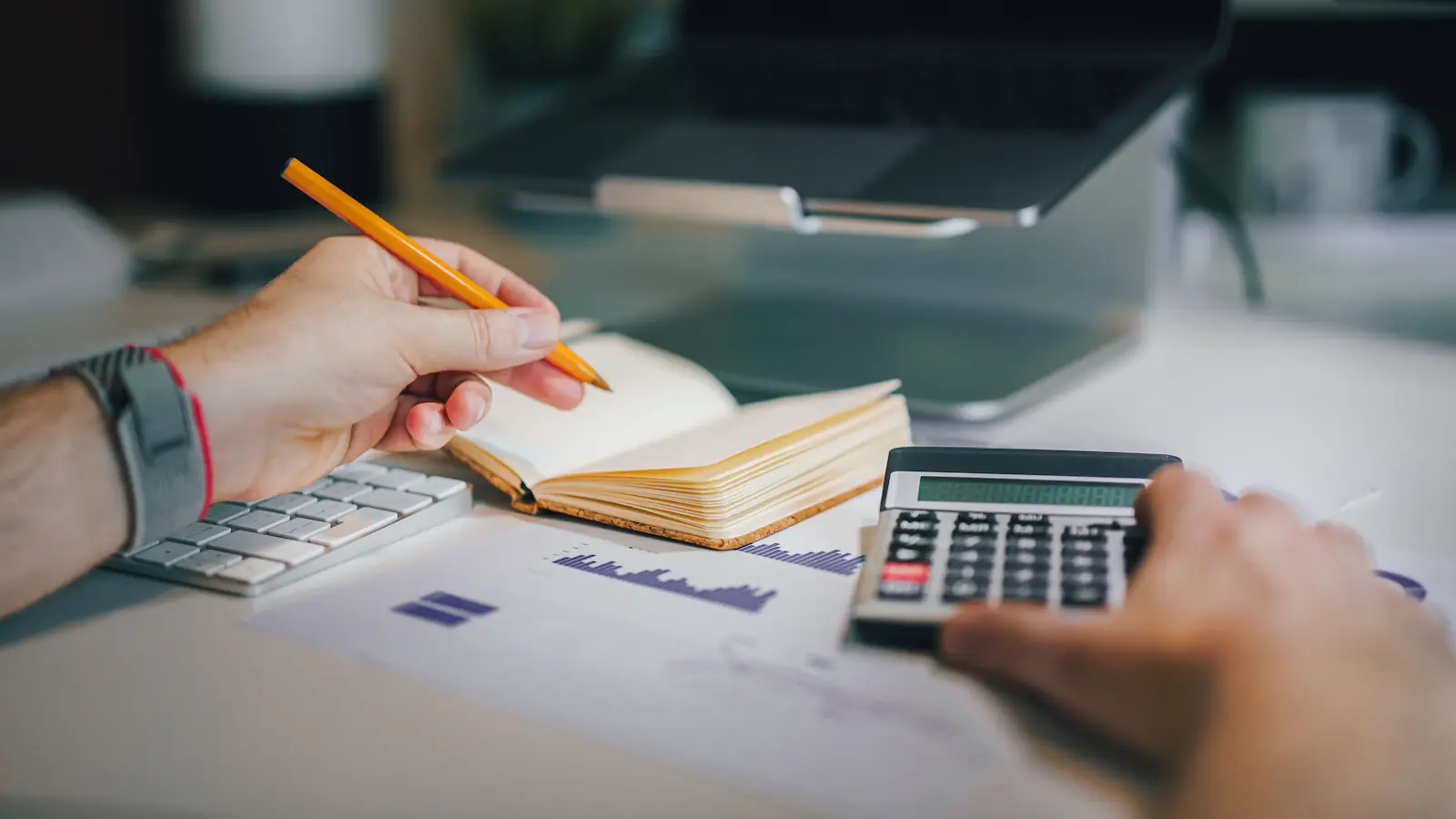 Hände mit Bleistift und Taschenrechner über einem Notizbuch und ausgedruckten Diagrammen