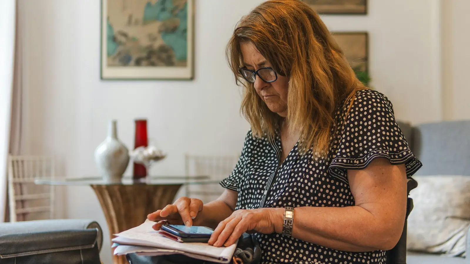 Eine Frau sitzt zu Hause am Tisch, schreibt auf ein Blatt Papier und sieht auf ihr Handy