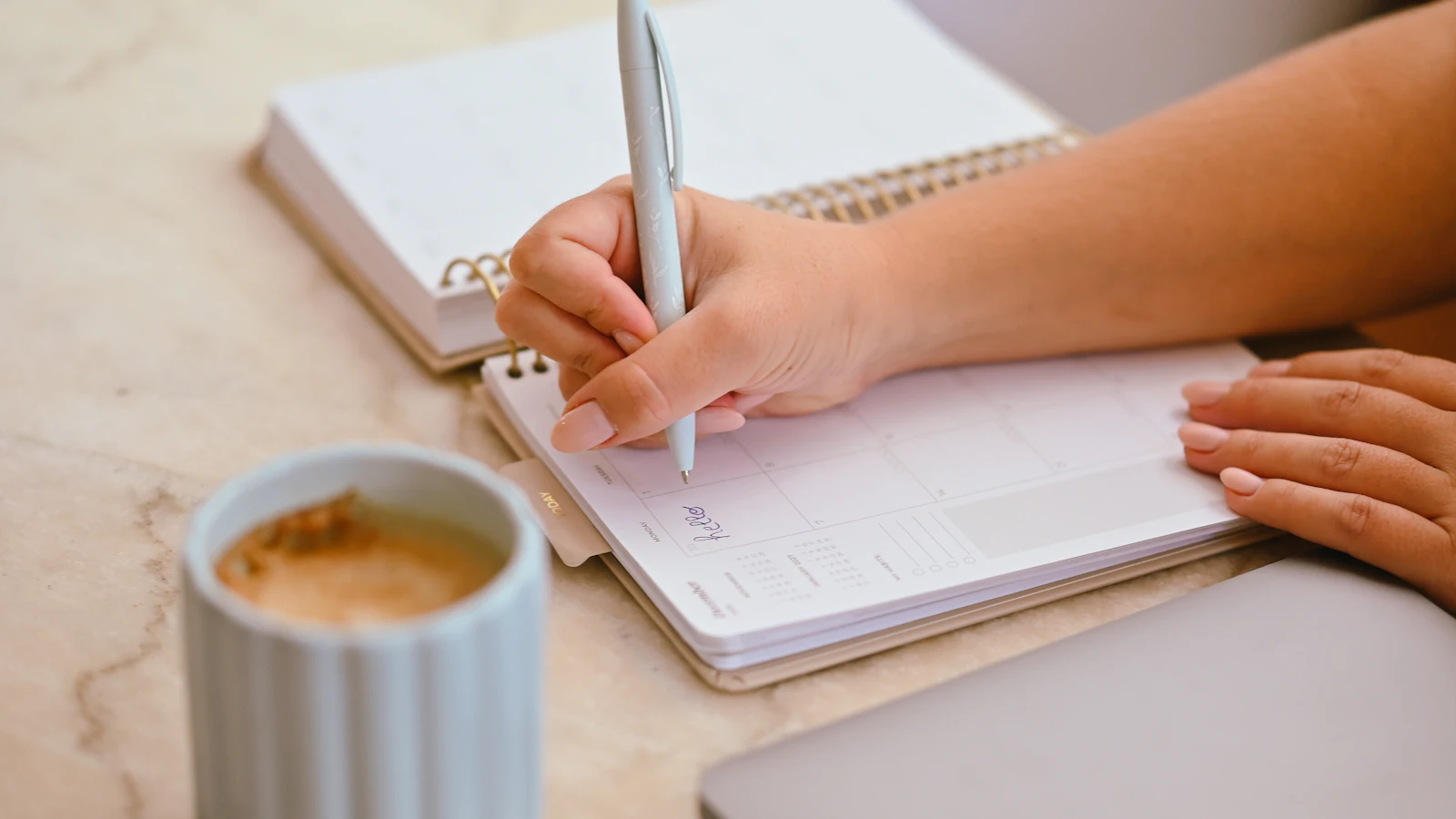 Eine Hand schreibt mit einem Stift in einen aufgeschlagenen Kalender, daneben eine Tasse Kaffee