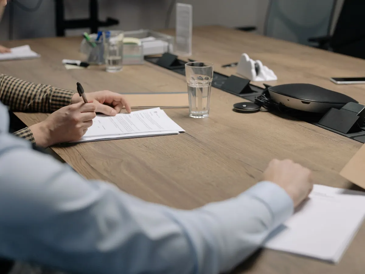 Besprechungstisch mit Unterlagen, eine Hand mit Stift setzt zur Unterschrift an