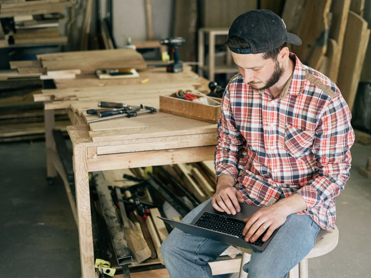 Ein Handwerker sitzt in seiner Werkstatt mit dem Laptop auf den Knien und bereitet den Jahresabschluss seiner UG selbst vor