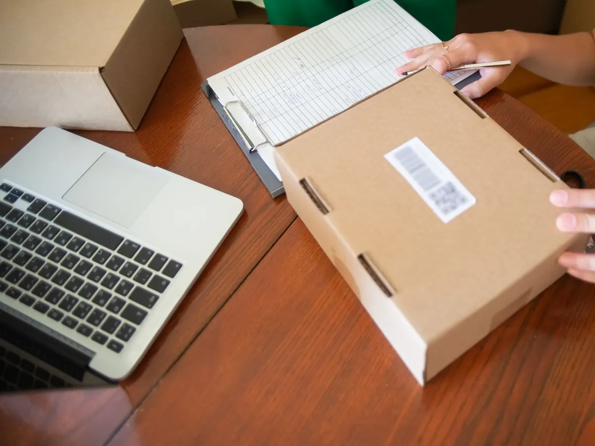 Laptop, Klemmbrett und ein Versandkarton mit Etikett auf dem Packtisch – im Onlinehandel entsteht jede Bestellung zuerst im Vorprogramm