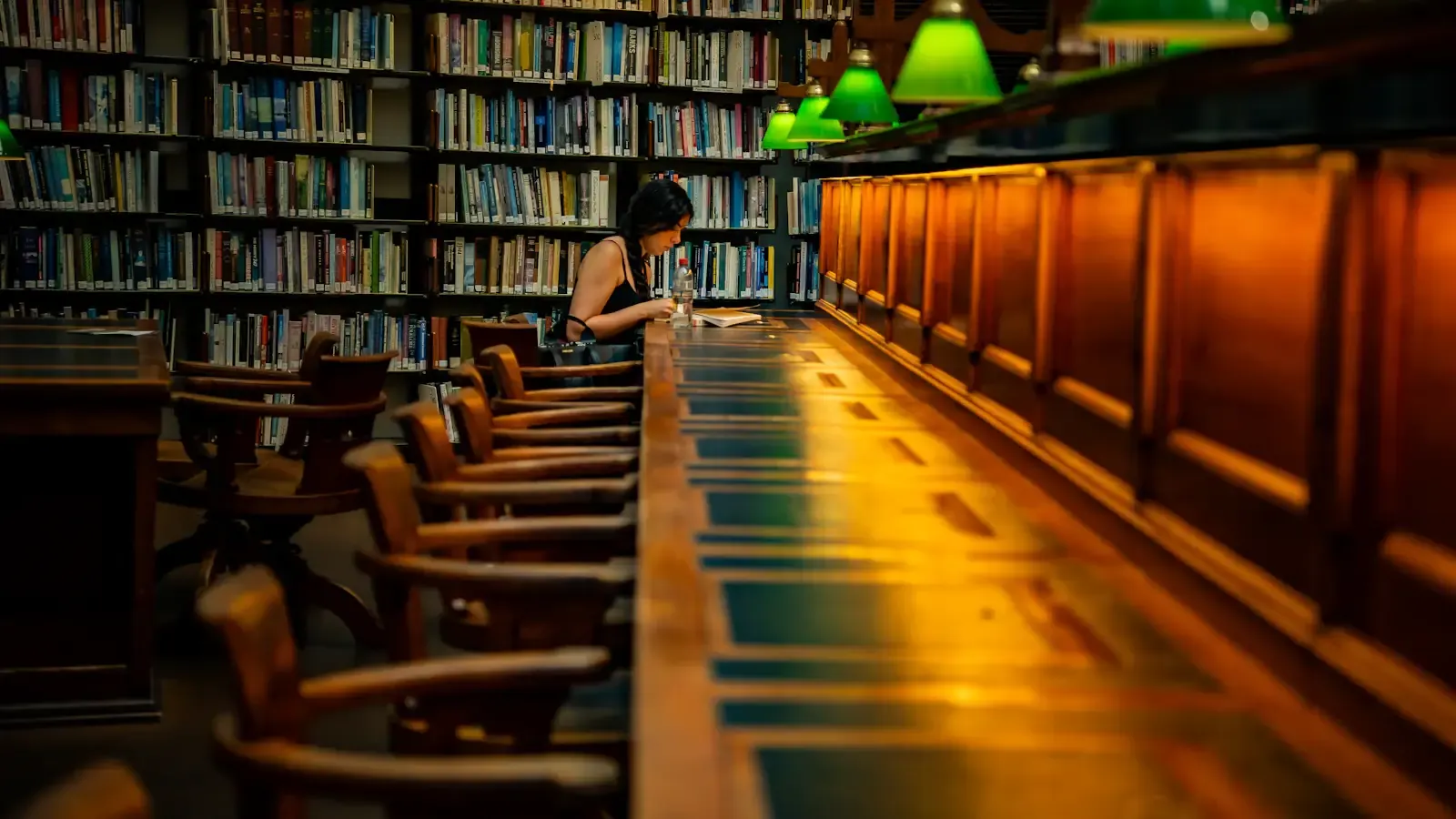 Eine Frau liest allein an einem langen Tisch in einer Bibliothek mit grünen Leselampen
