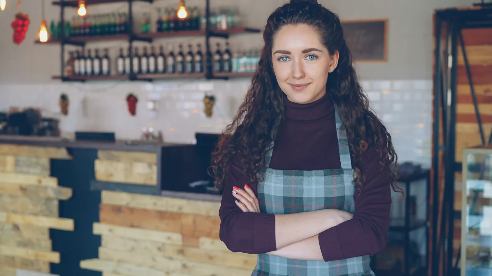 Eine Frau mit Schürze steht mit verschränkten Armen in einem kleinen Café vor der Theke