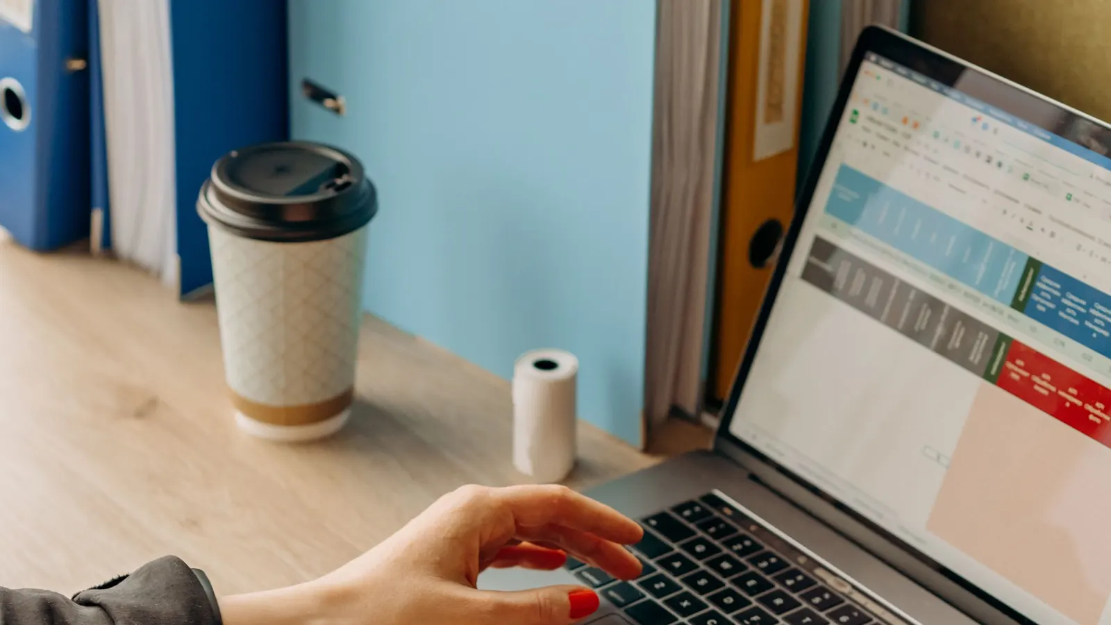 Hände tippen am Laptop, daneben Ordner und ein Kaffeebecher